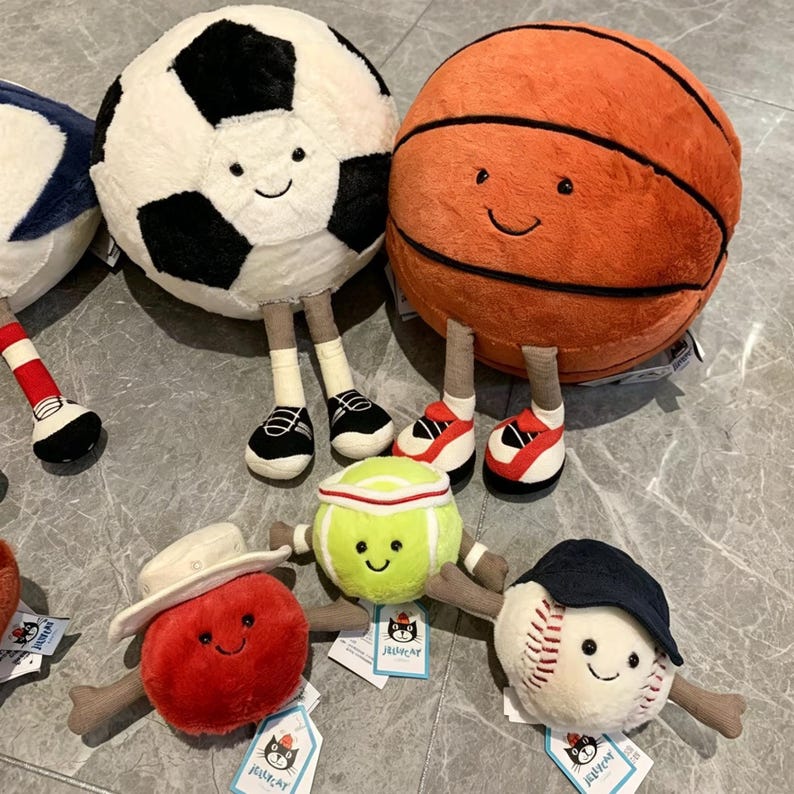 Puede incluir: Una colecci&oacute;n de pelotas deportivas de peluche con caras sonrientes y piernas adjuntas. Incluye un bal&oacute;n de f&uacute;tbol, un bal&oacute;n de baloncesto, una pelota de tenis, una pelota de b&eacute;isbol y una manzana roja. El bal&oacute;n de f&uacute;tbol tiene paneles en blanco y negro, y el bal&oacute;n de baloncesto es naranja.