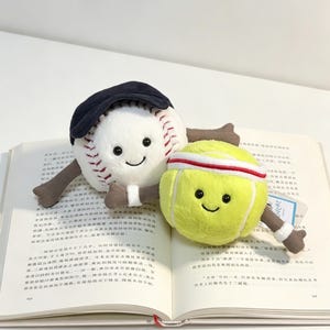 Puede incluir: Dos juguetes de peluche: una pelota de b&eacute;isbol con una gorra azul marino y una pelota de tenis con una cinta para la cabeza blanca y roja. Ambos tienen caras sonrientes y extremidades marrones. Est&aacute;n sobre un libro abierto.