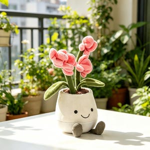May include: A plush toy flower arrangement featuring three pink flowers with green stems and leaves, planted in a white smiling pot with gray feet. The background shows a balcony with potted plants.