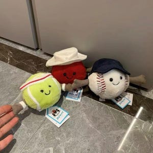 Puede incluir: Tres juguetes de peluche con forma de pelota deportiva: una pelota de tenis con una diadema blanca, una pelota roja con un sombrero blanco y una pelota de b&eacute;isbol con una gorra azul marino. Cada juguete tiene una cara sonriente y brazos adjuntos.