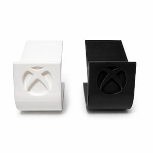 Peut inclure: Deux supports pour console de jeux, un blanc et un noir. Chaque support présente un design incurvé avec un emblème circulaire d'un 'X' stylisé au centre. Les supports sont conçus pour maintenir une console de jeux.