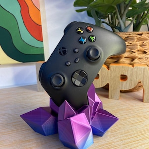 Pode incluir: Um comando de videojogo sem fios preto pousado num suporte impresso em 3D com a forma de um grupo de cristais geométricos. O suporte de cristal apresenta um esquema de cores degradé vibrante que vai do roxo escuro ao azul na base.