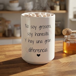 May include: A white ceramic mug filled with hot chocolate and marshmallows. The mug has black text that reads "No soy grosera, soy honesta. Y hay una gran diferencia" with a small black heart. A jar of honey and a honey dipper are in the background.