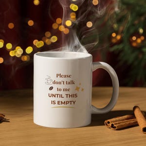 May include: White ceramic coffee mug with the text "Please don't talk to me UNTIL THIS IS EMPTY" in brown. A coffee bean and coffee cup graphic are also on the mug. Steam rises from the mug, suggesting a hot beverage.