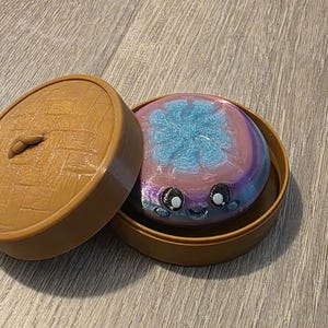 Könnte beinhalten: Ein brauner runder Behälter mit Deckel, der eine farbenfrohe Badekugel enthält. Die Badekugel ist eine Mischung aus Blau, Rosa und Lila mit einem niedlichen Gesichtsdesign. Der Deckel liegt daneben.