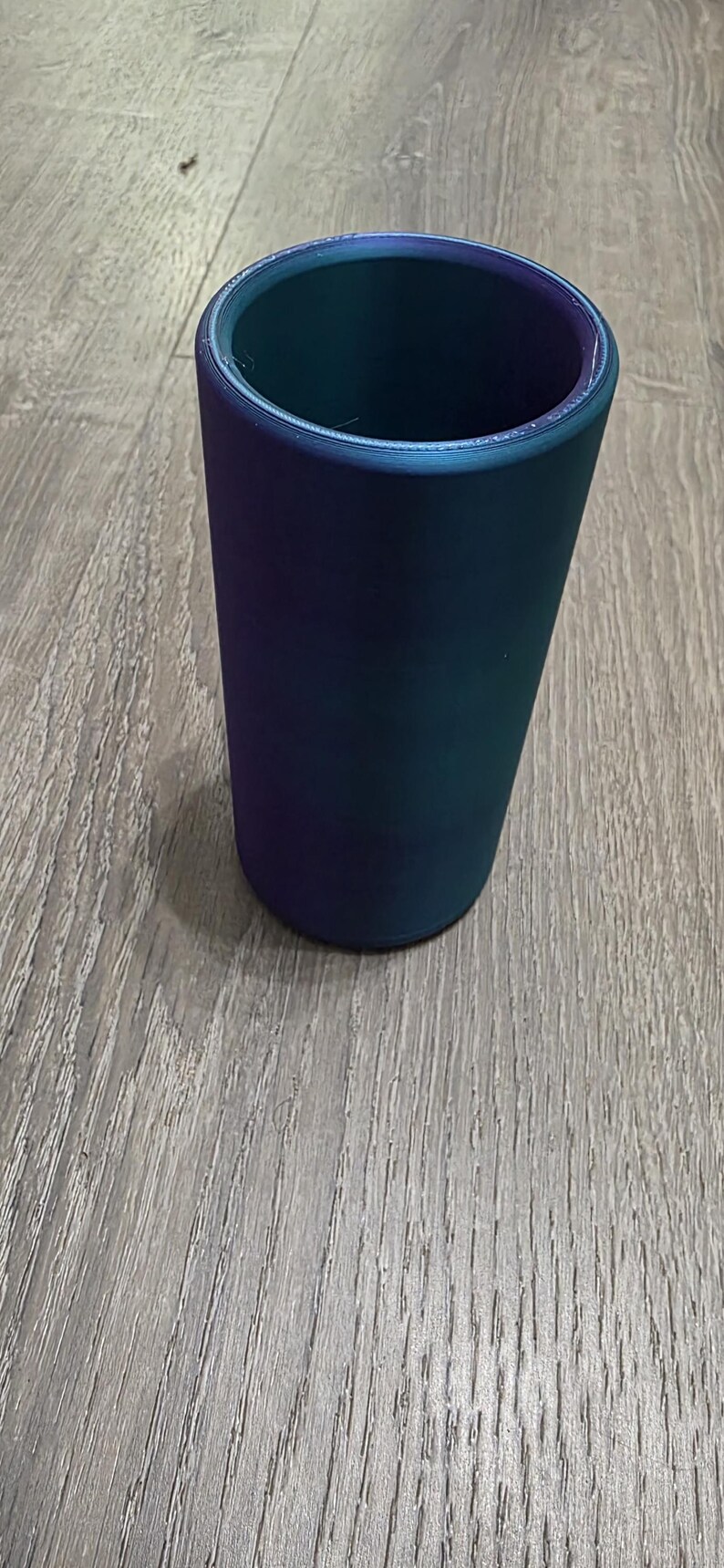 Puede incluir: Un vaso cil&iacute;ndrico con un exterior liso y s&oacute;lido que presenta un acabado de cambio de color que transita entre tonos profundos de p&uacute;rpura, azul y verde azulado. El recipiente tiene un dise&ntilde;o simple de boca abierta y una base plana.