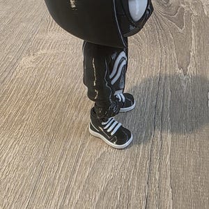 Puede incluir: Una figura negra de un personaje con una cabeza grande, que lleva una chaqueta con capucha, pantalones y zapatillas deportivas. La figura tiene ojos blancos y un dise&ntilde;o blanco en la chaqueta y los pantalones. Las zapatillas son negras con rayas blancas.