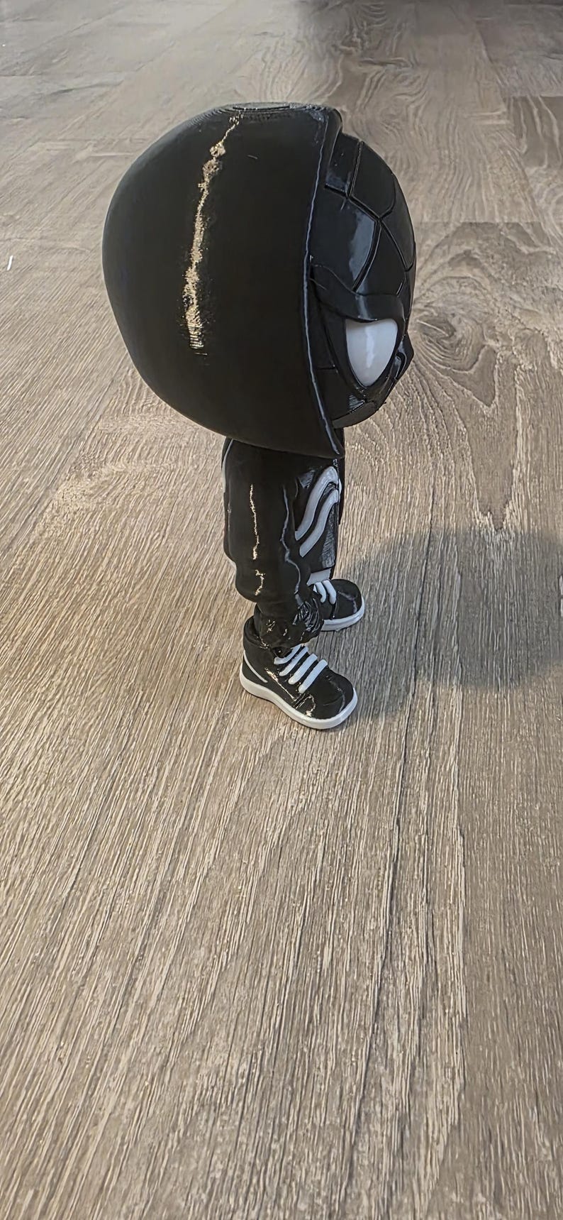 Puede incluir: Una figura negra de un personaje con una cabeza grande, que lleva una chaqueta con capucha, pantalones y zapatillas deportivas. La figura tiene ojos blancos y un dise&ntilde;o blanco en la chaqueta y los pantalones. Las zapatillas son negras con rayas blancas.