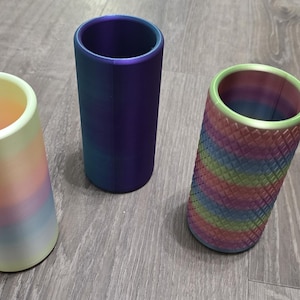 Puede incluir: Tres soportes cil&iacute;ndricos dispuestos verticalmente. El soporte de la izquierda presenta un degradado pastel suave de amarillo, rosa y azul. El soporte central es de un color violeta met&aacute;lico s&oacute;lido y profundo. El de la derecha tiene un patr&oacute;n de rombos texturizado en colores arco&iacute;ris.