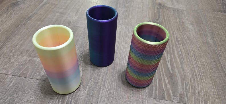 Puede incluir: Tres soportes cil&iacute;ndricos dispuestos verticalmente. El soporte de la izquierda presenta un degradado pastel suave de amarillo, rosa y azul. El soporte central es de un color violeta met&aacute;lico s&oacute;lido y profundo. El de la derecha tiene un patr&oacute;n de rombos texturizado en colores arco&iacute;ris.