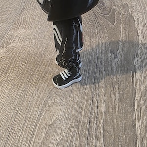 Puede incluir: Una figura negra de un personaje con una cabeza grande, que lleva un atuendo negro con rayas blancas y zapatillas blancas. La figura tiene una m&aacute;scara blanca y est&aacute; de pie sobre una superficie de madera.