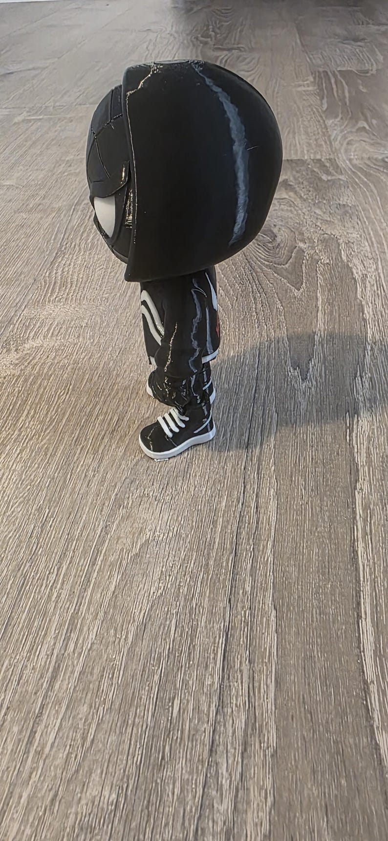 Puede incluir: Una figura negra de un personaje con una cabeza grande, que lleva un atuendo negro con rayas blancas y zapatillas blancas. La figura tiene una m&aacute;scara blanca y est&aacute; de pie sobre una superficie de madera.