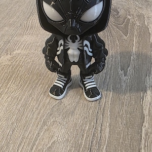 Puede incluir: Una figura Funko Pop de Spider-Man en blanco y negro. La figura lleva una sudadera con capucha y una chaqueta negras con un emblema de ara&ntilde;a blanco. Tiene ojos blancos y lleva zapatillas deportivas en blanco y negro.