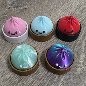 May include: Five colorful, dumpling-shaped objects in small bamboo steamers. The dumplings are pink, blue, red, green, and purple, each with a smiling face. The steamers are brown, black, and white.