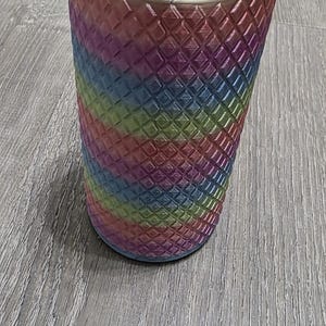 Puede incluir: Un vaso cil&iacute;ndrico con un acabado degradado de arco&iacute;ris vibrante que transita por tonos verdes, azules, morados y rosas. La superficie exterior est&aacute; cubierta con una textura de rejilla en forma de diamante en relieve.