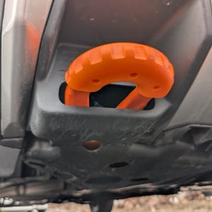 May include: Close-up of a vehicle's orange tow hook, a U-shaped component with a textured grip. The hook is set within a black housing, mounted on the vehicle's underside. The vehicle's bumper and undercarriage are visible.