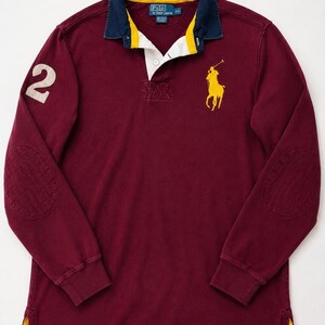 May include: A long-sleeve burgundy polo shirt featuring a navy blue collar with yellow trim, a white button placket, and a yellow embroidered polo player logo on the chest. The right sleeve displays a large white number 2, and both elbows have patches.