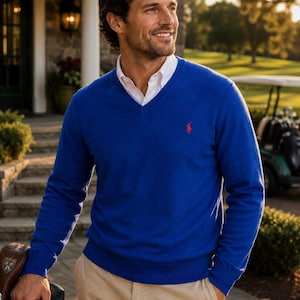 May include: A bright blue V-neck sweater featuring a small red embroidered logo on the left chest, worn over a crisp white collared shirt, paired with light tan trousers. The sweater has long sleeves with ribbed cuffs and a ribbed hemline.