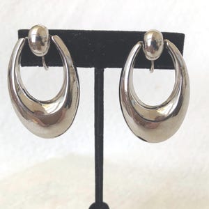 Puede incluir: Un par de pendientes de clip ovalados plateados. Cada pendiente tiene una superficie lisa y pulida y un pequeño clip redondo en la parte superior. Los pendientes se exhiben en un soporte de pendientes de terciopelo negro sobre un fondo blanco.