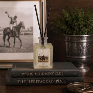 May include: A square glass reed diffuser bottle filled with light-colored liquid and three black reeds. The label features a polo match illustration and the text "THE POLO CLUB". It rests on two stacked books titled "BEYOND THE PONY CLUB" and "THE HERITAGE OF POLO".