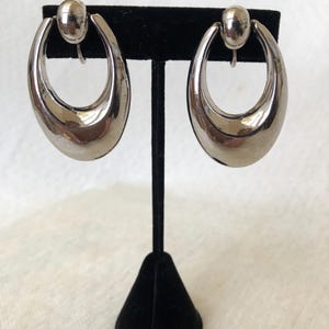 Puede incluir: Un par de pendientes de clip ovalados plateados. Cada pendiente tiene una parte superior redondeada y una superficie lisa y pulida. Los pendientes se exhiben en un soporte de pendientes de terciopelo negro sobre un fondo blanco.