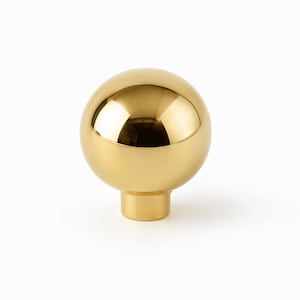 Op de afbeelding: Gouden meubelknoppunt in de vorm van een bol met een gepolijste afwerking. De ronde bal zit op een korte, cilindrische basis. Het reflecterende oppervlak creëert highlights en schaduwen.