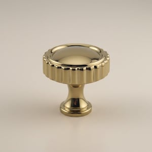 May include: A gold-colored cabinet knob with a fluted edge and a rounded top. The knob has a pedestal base and a shiny, reflective surface. This decorative hardware is suitable for drawers and cabinets.