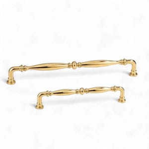 May include: Two gold-toned metal cabinet pulls with a polished finish. Each handle features a central spherical accent, tapered cylindrical bars, and curved ends that attach to a mounting surface with circular bases for a secure installation.