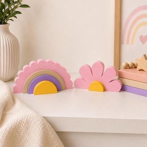 K&ouml;nnte beinhalten: Zwei dekorative Holzst&uuml;cke: ein rosa Regenbogen mit gelben, lila und beigen Schichten und eine rosa Blume mit gelber Mitte. Sie stehen auf einer wei&szlig;en Oberfl&auml;che, mit einem gerahmten Regenbogen-Druck im Hintergrund. Eine wei&szlig;e Vase mit Gr&uuml;nzeug befindet sich links.