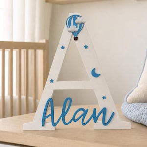 May include: A white wooden letter "A" with blue accents, featuring a hot air balloon and stars. The name "Alan" is written in blue script. Decorative item for a nursery or child's room.