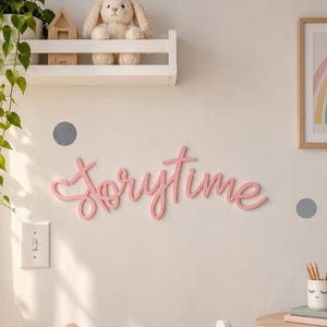 May include: A pink script wall sign reading "storytime" mounted on a wall, accompanied by grey and pink circular wall decals. The scene includes a white shelf with a plush rabbit, a woven basket with a pink heart pillow, and a wooden chair.
