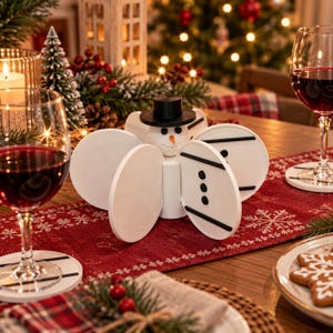 May include: A white snowman-shaped coaster holder with a black top hat, orange carrot nose, and black button details. The holder features four circular slots for storing white coasters, each decorated with two horizontal black lines and three black dots.