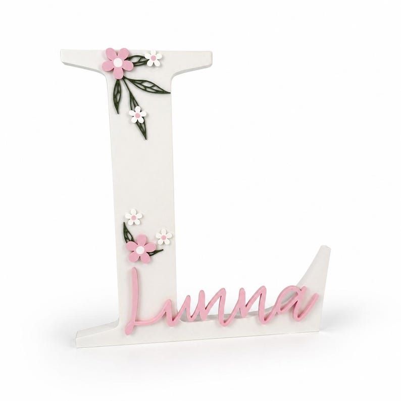 May include: A white wooden letter 'L' adorned with pink and white floral embellishments and green leaves. The name 'Luna' is written in pink cursive script. The decorative letter is designed to stand upright, suitable for a nursery or bedroom.