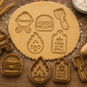 May include: A set of four brown plastic cookie cutters featuring a grill, a hamburger, a flame shape with the text DAD, and a rectangular plaque with the text KING OF THE GRILL. The cutters are shown alongside impressions made in cookie dough.