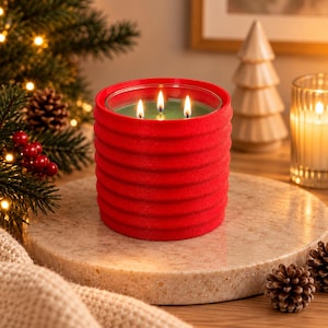 May include: A three-wick candle in a cylindrical red container with a textured, ribbed exterior. The wax inside is green, and the three wicks are lit, producing small, bright flames. The candle sits upright, showcasing its vibrant red color.