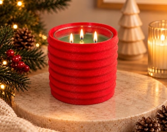 May include: A three-wick candle in a cylindrical red container with a textured, ribbed exterior. The wax inside is green, and the three wicks are lit, producing small, bright flames. The candle sits upright, showcasing its vibrant red color.