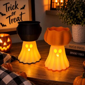 May include: Two illuminated ghost-shaped candle holders, one topped with a black cauldron and the other with an orange pumpkin bowl. Both ghosts feature cut-out eyes and mouths that glow with a warm light, creating a festive Halloween display.