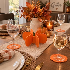 May include: A set of bright orange silicone pumpkin-shaped coasters placed on a wooden dining table. Each coaster features a scalloped edge and a flat surface designed to hold a wine glass. The coasters are arranged around a centrepiece.