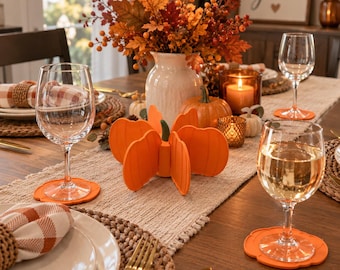 May include: A set of bright orange silicone pumpkin-shaped coasters placed on a wooden dining table. Each coaster features a scalloped edge and a flat surface designed to hold a wine glass. The coasters are arranged around a centerpiece.