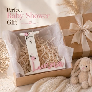 May include: A white wooden letter "L" with pink floral accents and the name "Luna" in pink, presented in a gift box with shredded paper. A plush bunny and a wrapped gift box are also visible.