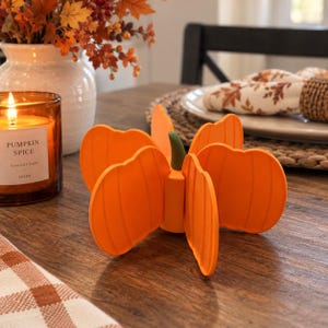 May include: A bright orange pumpkin-shaped trivet featuring a segmented design with vertical ridges and a small green stem at the centre. The object is constructed from a flexible material and stands upright on a flat surface, displaying a symmetrical form.
