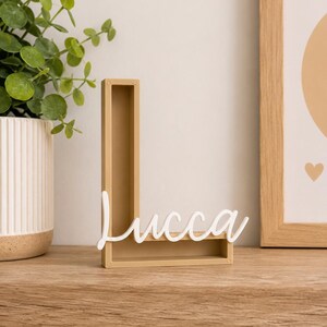 May include: A tan, hollow, three-dimensional letter L made of a solid material. Attached to the front of the letter is a white, cursive script cutout of the name Lucca. The object stands upright on a flat surface, displaying a clean, modern design.