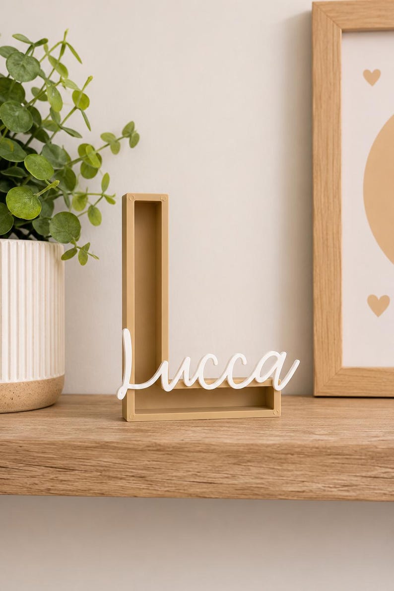 May include: A tan, hollow, three-dimensional letter L made of a solid material. Attached to the front of the letter is a white, cursive script cutout of the name Lucca. The object stands upright on a flat surface, displaying a clean, modern design.