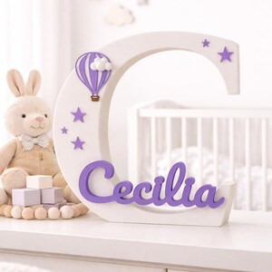 May include: A white wooden letter "C" with purple accents, featuring a hot air balloon, stars, and the name "Cecilia." A plush bunny and wooden blocks are in the background. The letter is approximately 12 inches tall.