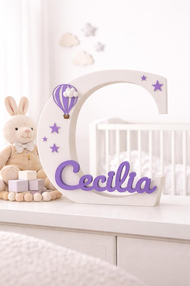 May include: A white wooden letter "C" with purple accents, featuring a hot air balloon, stars, and the name "Cecilia." A plush bunny and wooden blocks are in the background. The letter is approximately 12 inches tall.