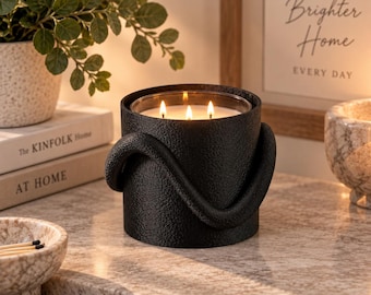 Puede incluir: Un portavelas cilíndrico negro con textura, que presenta un asa decorativa curva en forma de serpiente que rodea el exterior. La vela del interior tiene tres mechas encendidas, proyectando un brillo cálido desde el centro del recipiente mate.