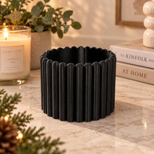 May include: A cylindrical black planter featuring a vertical fluted texture along its exterior surface. The container has a scalloped top edge and a hollow interior, designed with a consistent matte black finish throughout its circular structure.
