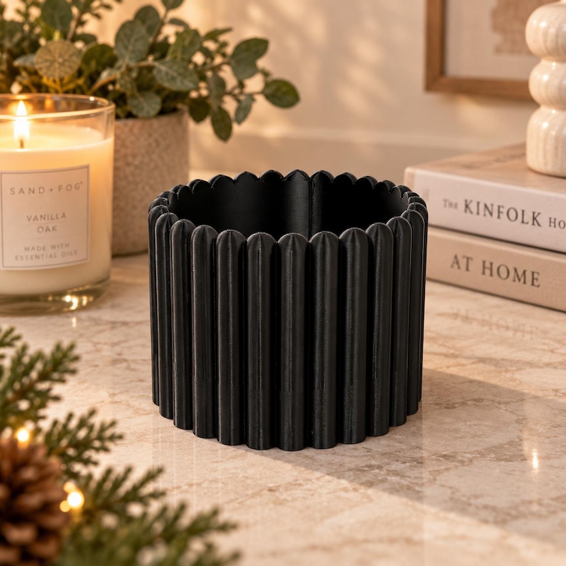 May include: A cylindrical black planter featuring a vertical fluted texture along its exterior surface. The container has a scalloped top edge and a hollow interior, designed with a consistent matte black finish throughout its circular structure.
