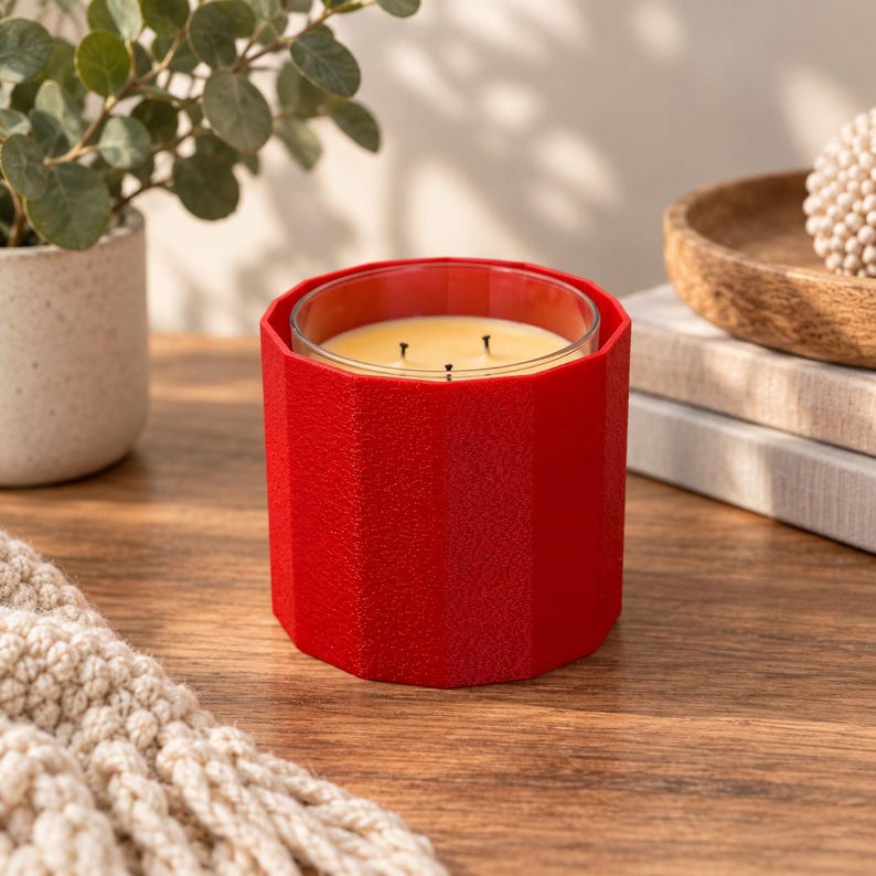 May include: A three-wick candle in a clear glass container, encased in a vibrant red, faceted, textured sleeve. The candle wax is a pale yellow color. The sleeve features a geometric, multi-sided design that surrounds the glass vessel entirely.