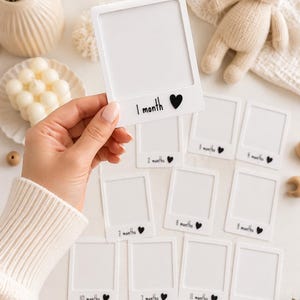 May include: A set of white, rectangular photo frames designed to look like instant film prints. Each frame features a black printed label indicating a month from 1 month to one year, accompanied by a small black heart icon next to the text.